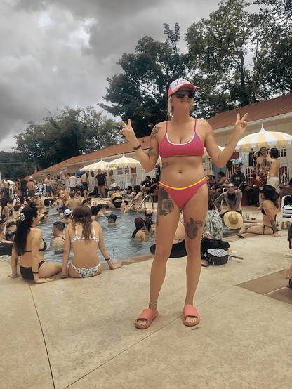 A woman wearing a pink and yellow bikini stands in the center of a crowded outdoor swimming pool area, making peace signs with both hands. The background shows other people in the pool, umbrellas, and buildings under a cloudy sky. I PEE IN POOLS HAT