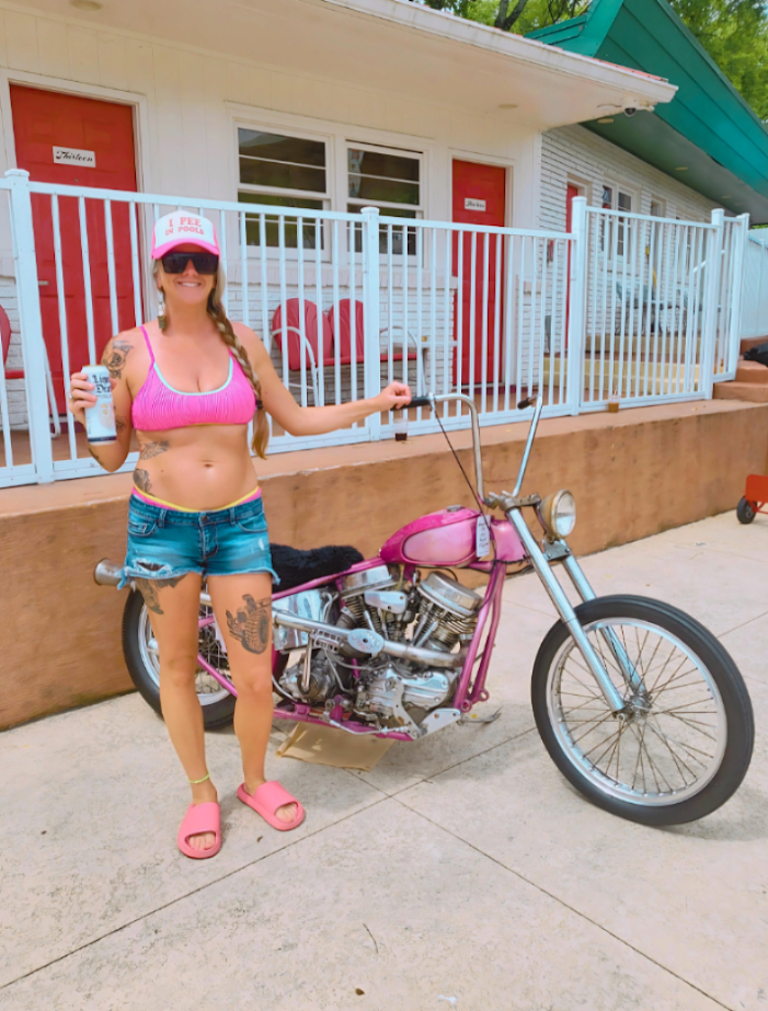 A woman stands next to a custom pink chopper motorcycle in front of a motel-style building with red doors and a white railing. She is wearing a pink bikini top, denim shorts, pink sandals, and a pink I PEE IN POOLS cap, holding a drink.